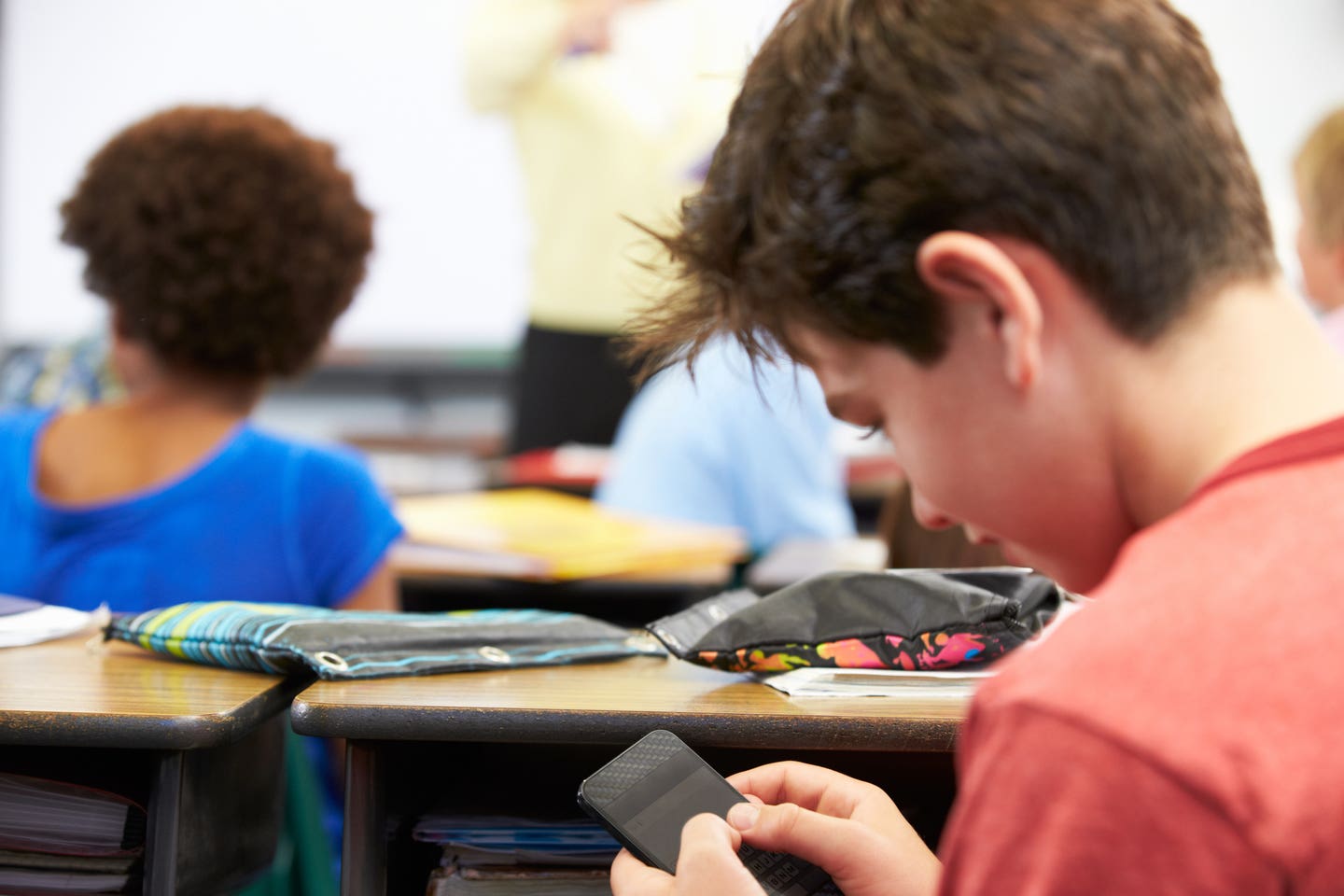 Pupil Sending Text Message On Mobile Phone In Class Sitting At Desk